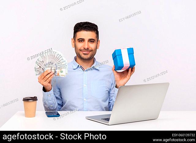 Purchase of holiday gifts, internet shopping. Happy elegant man sitting workplace with laptop, holding money dollars and present box, smiling at camera