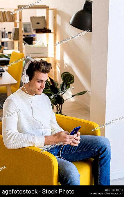 Young man with smart phone listening music through headphones in living room of new home