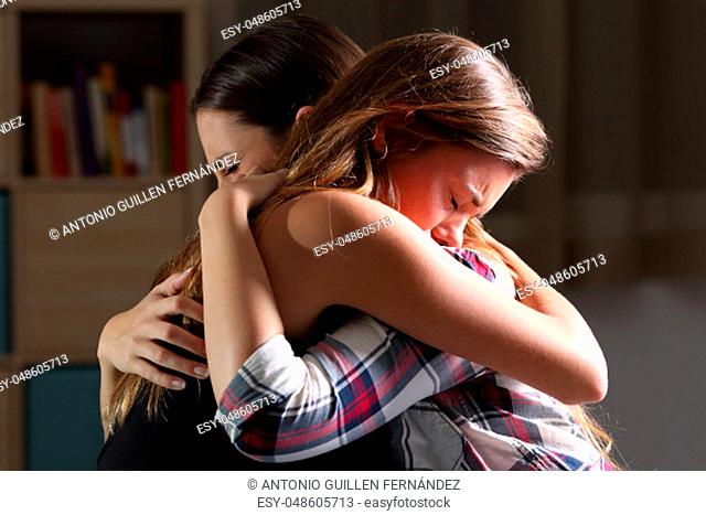 Side view of two sad good friends embracing in a bedroom in a house interior with a dark light in the background