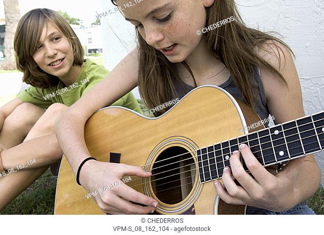 Close-up of a teenage girl playing the guitar with a teenage boy sitting beside her