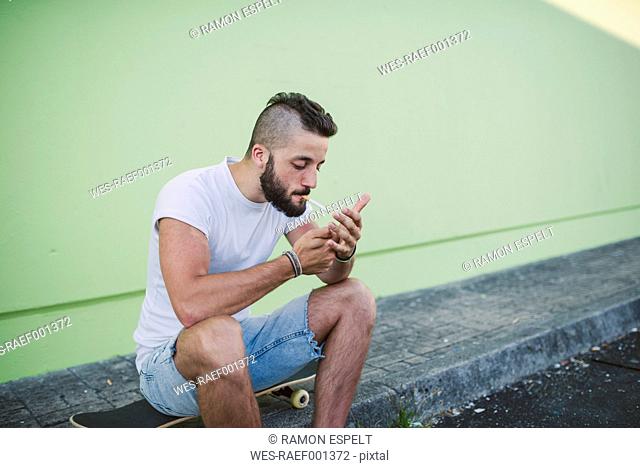 Young man sitting on skateboard, lighting a cigarette
