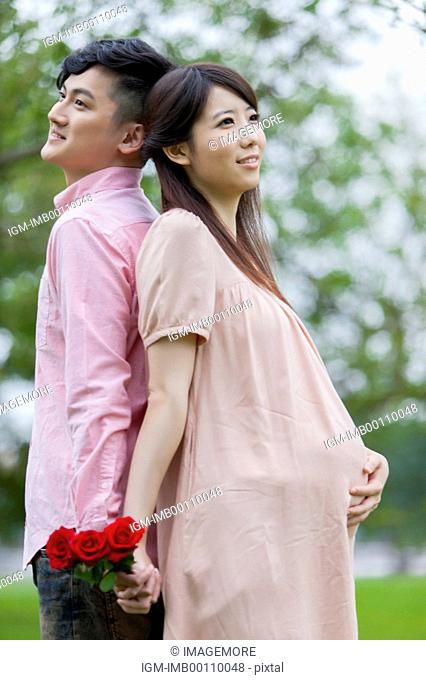 Young man and pregnant woman standing with back to back and holding hands with smile