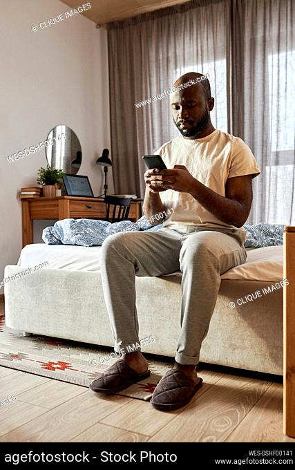 Man using smart phone sitting on bed in morning at home