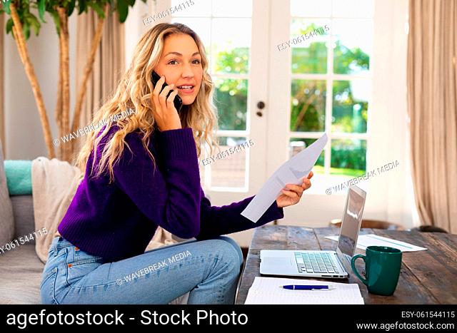 Smiling caucasian woman using laptop, talking on smartphone and holding paperwork working at home