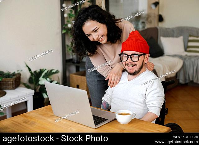 Smiling woman standing by boyfriend in wheelchair looking at laptop