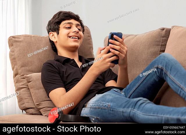 A CHEERFUL TEENAGER RESTING ON SOFA AND USING MOBILE PHONE