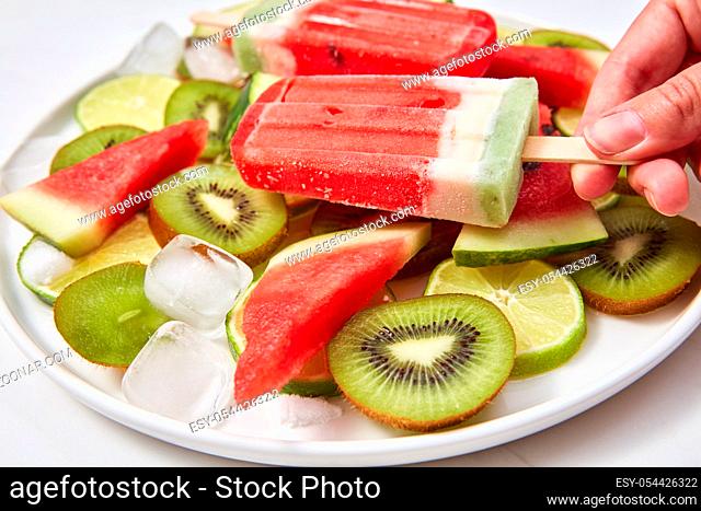 A woman's hand holds an ice cream on a stick in the form of a piece of watermelon on a background of a plate with pieces of different fruits around a white...
