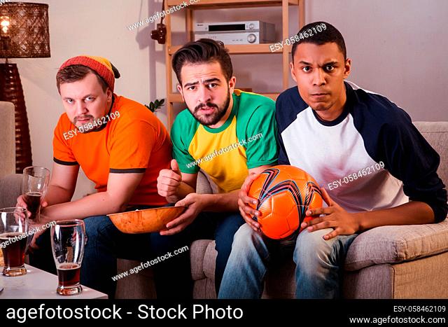 Picture of serious friends watching football game while sitting on sofa or couch and looking at camera. Football concept