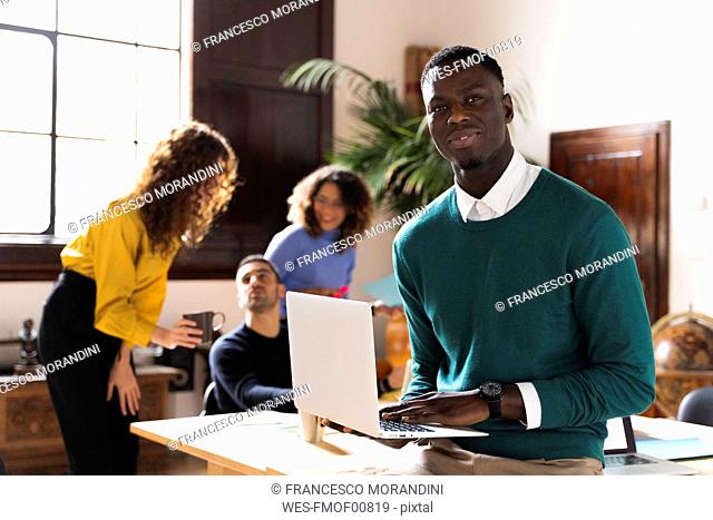 Portrait of a confident man in office using laptop with colleagues in background