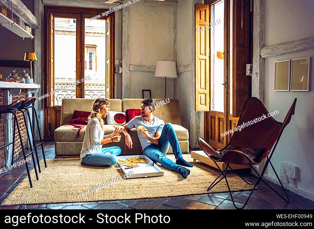 Young couple sitting on floor of living room and eating pizza from box