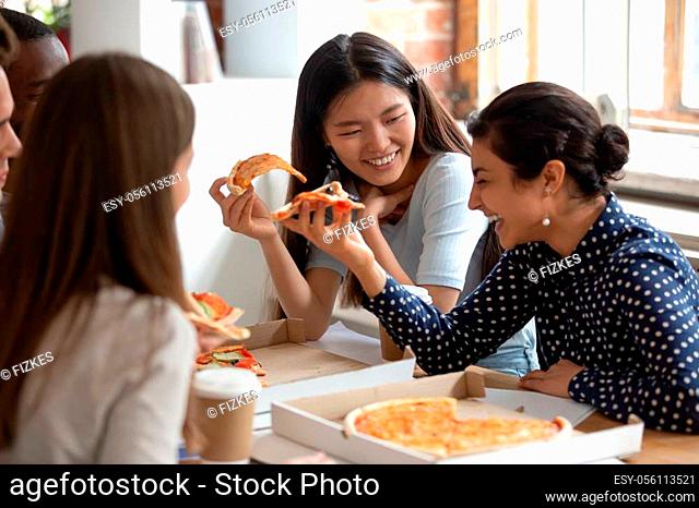 Happy multiracial friends diverse students schoolmates sitting at table indoors eat share pizza together during lunch, pretty girls and guys having fun