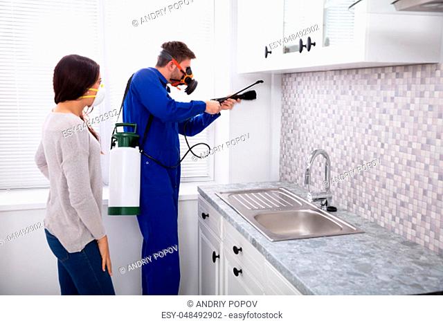Pest Control Worker And Woman Spraying Pesticide With Torch In Kitchen