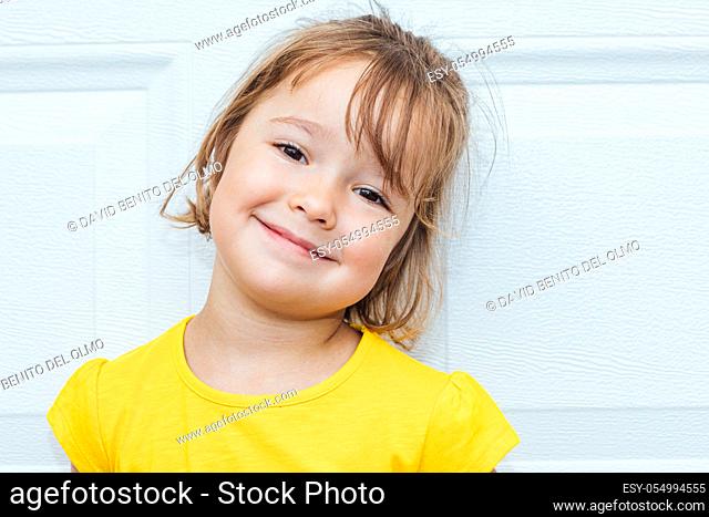 Adorable blond-haired girl wearing a yellow shirt leaning against white background