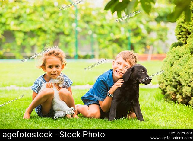 Caring brother and sister embracing kitten and puppy on green lawn. Cute little girl holding white kitten, smiling schoolboy embracing black puppy