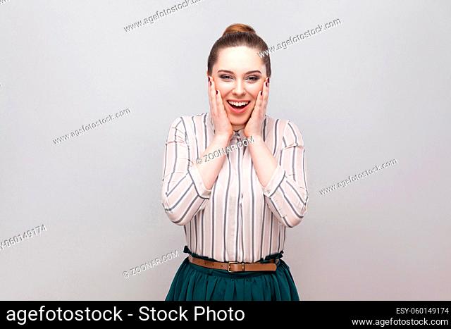 Portrait of happy surprised beautiful young woman in striped shirt, collected ban hairstyle, standing, amazed and looking at camera with toothy smile