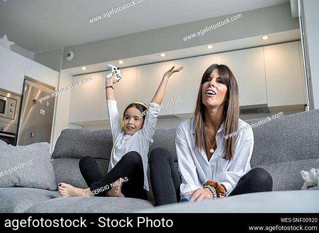 Happy daughter with hand raised sitting by mother while playing video games at home