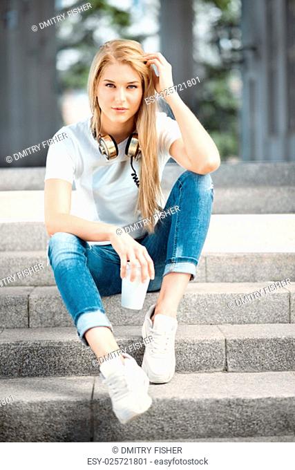 Happy young woman wearing vintage music headphones around her neck, sitting on stairs with a take away coffee cup and looking straight against urban city...