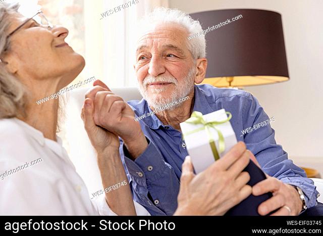 Senior man looking at woman sitting with gift at hotel apartment