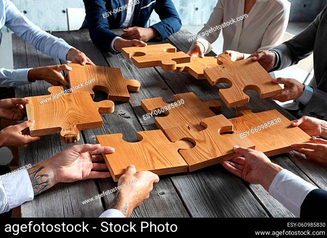 Business people team sitting around meeting table and assembling wooden jigsaw puzzle pieces unity cooperation ideas concept