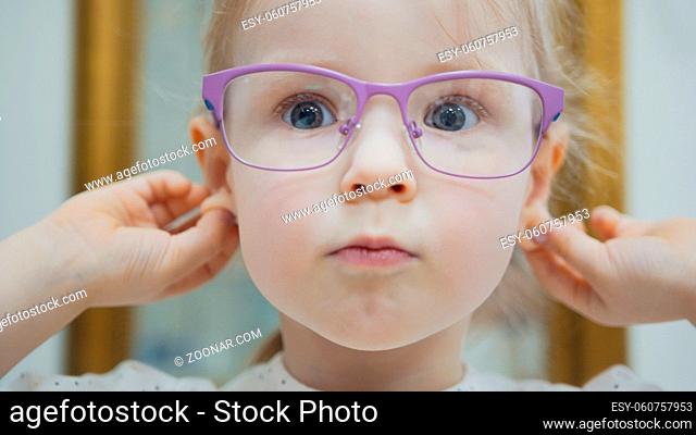 Little girl tries fashion medical glasses near mirror - shopping in ophthalmology clinic, close up