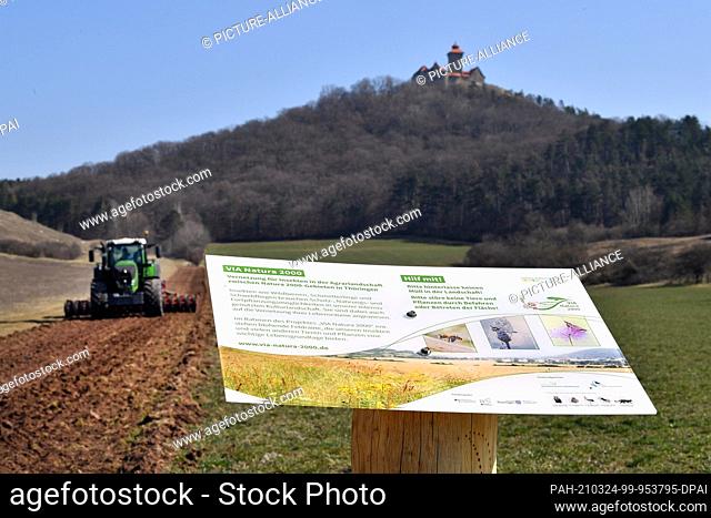 24 March 2021, Thuringia, Mühlberg: A board informs about a nature conservation measure, while a tractor with a seed drill drives over a field not far from...
