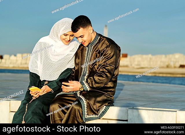 Young woman sitting by man using smart phone on sunny day