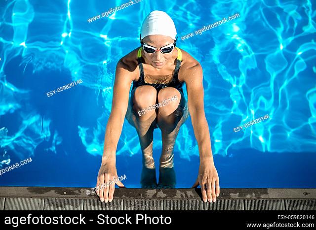 Charming swimmer in the swimming pool. Woman wears a black-lime swimsuit, a white swim cap and swim glasses. She looks forward and holds her hands on the pool...