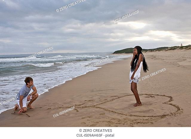 Young man on one knee on beach, young woman standing in heart shape