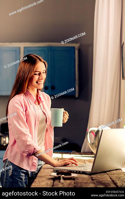 Smiling girl at home standing near window holding cup. Happy female in the kitchen looking at laptop, drinking tea or coffee