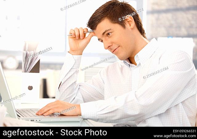 Young office worker thinking, sitting at office desk holding pen, using laptop computer, looking at screen, smiling