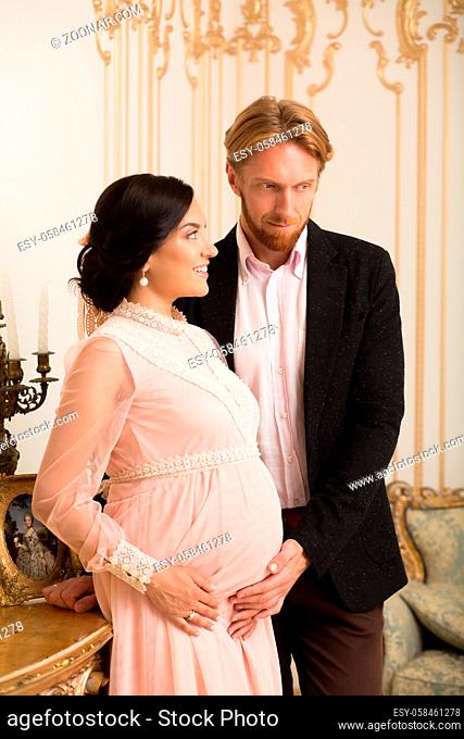 Portrait of handsome blond businessman touching his wife's belly. Brunette lady in pink dress expecting baby soon. Baby in royal family concept