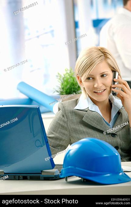 Smiling architect talking on phone looking at computer screen in office