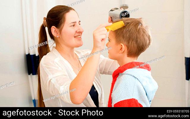 Portrait of young smiling mother drying and brushing wet hair of her little son after washing in bath. Concept of child hygiene and health care at home