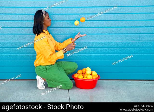 Young woman juggling with oranges in front of blue roller shutter