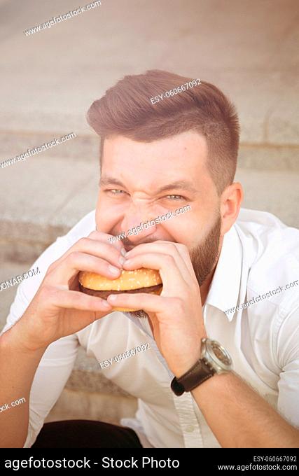City hipster businessman eating hamburger. Close-up portrait of hungry man having a snack during his break after hard work. Toned image