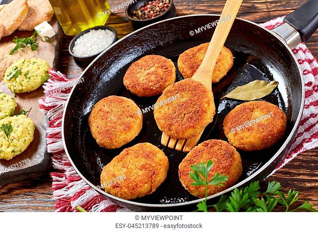 crispy rice cutlets of leftover rice with crumbled cheese and greens on a skillet on wooden table with uncooked cutlets, paneer cheese cubes on cutting board