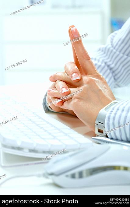 Closeup of female hands with fingers crossed at desk beside computer keyboard