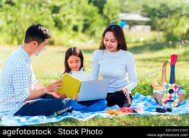 Happy Asian lifestyle young family father, mother and little girl having fun outdoor sitting on picnic blanket using laptop computer technologies while relaxing...