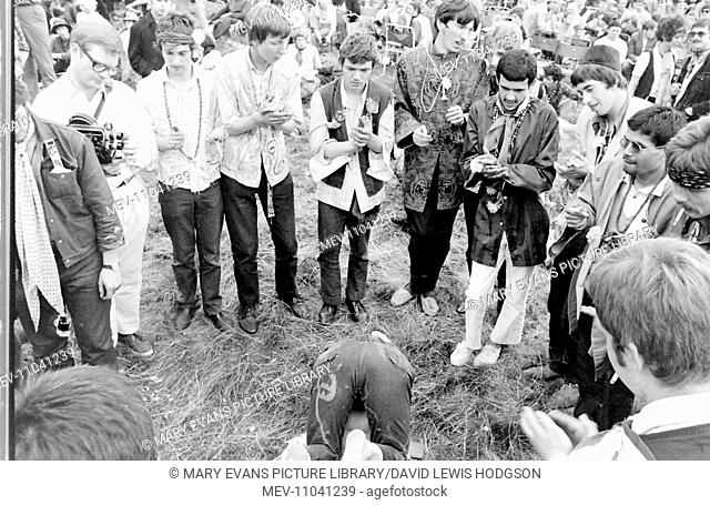 A group of hippies in a field at a Love-In at Woburn Park, Bedfordshire, standing and watching while a man prepares to do a headstand