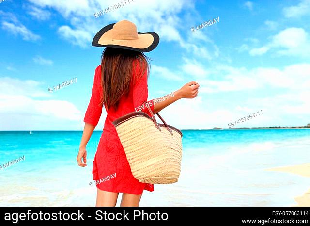 Woman tourist walking on tropical summer vacation wearing sun hat, red dress and beach bag relaxing on travel holidays. Young lady from behind in luxury fashion...