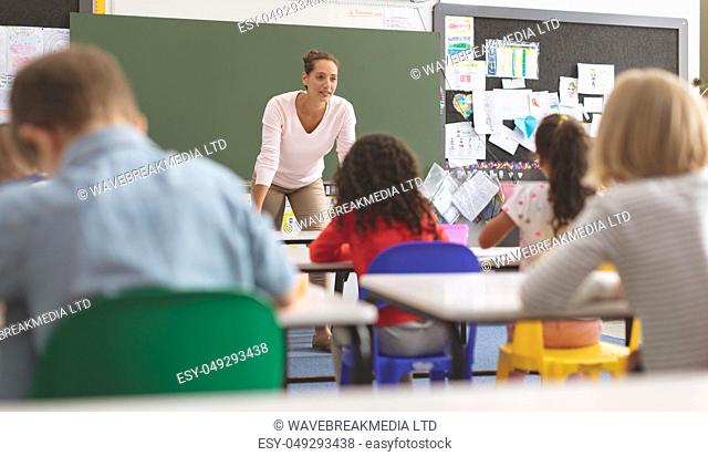 Front view of a teacher talking to school kids sitting on their chair in front of her against a greenboard in background