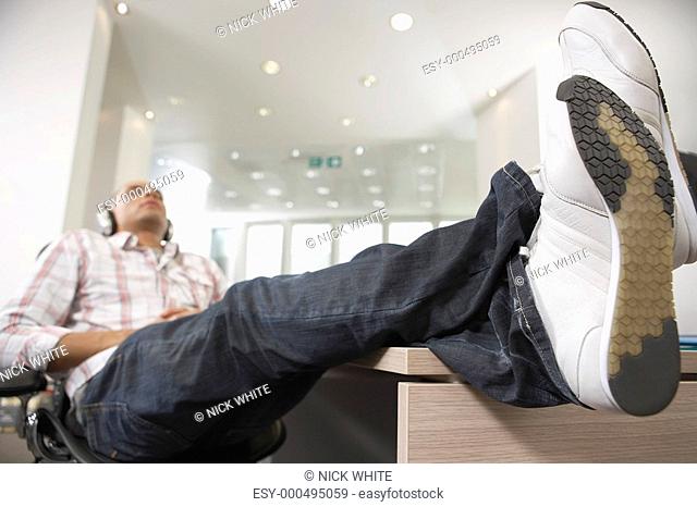 Casually dressed Businessman reclining with feet on desk Listening to Music With Headphones