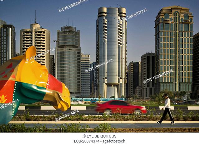 City centre skyline along the Corniche. Statue of a colourful patchwork camel, red car on road and a pedestrian