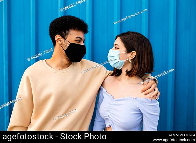Young couple wearing protective face mask looking at each other while standing against blue wall