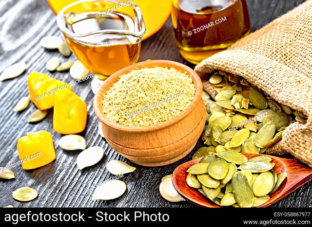 Pumpkin flour in a bowl, oil in a sauceboat and a jar, seeds in bag, on table and in spoon, slices of vegetable on dark wooden board background
