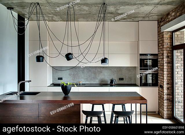 Kitchen in a loft style with concrete and brick walls. There is a kitchen island with a sink, plant and black chairs, light lockers with built-in oven and...