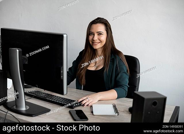 Brunette woman is working in front of a monitor in a office. Business woman working at computer at coworking space. Portrait of happy lady typing at workplace