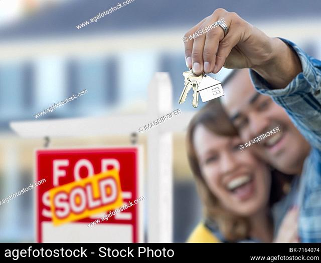 mixed-race couple in front of sold real estate sign and house with keys
