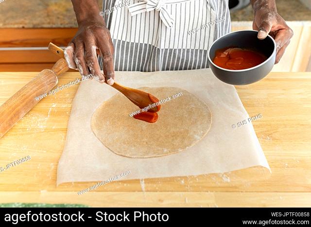 Man putting tomato sauce on pizza dough in kitchen at home
