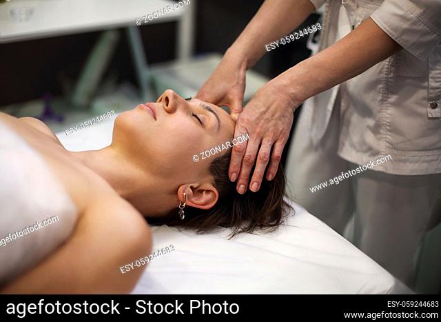 Unrecognizable masseuse in gown standing in spa salon and massaging face of female customer